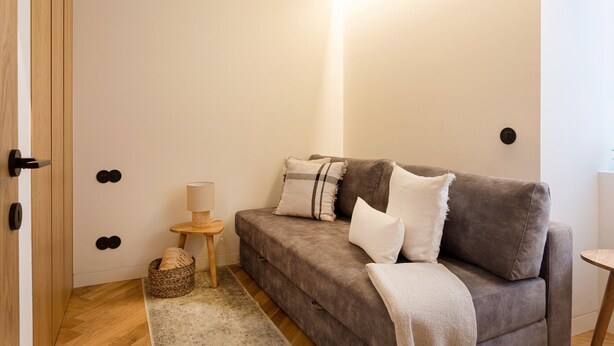 Ein Zimmer mit einer grauen Couch, einem kleinen runden Tisch, einer Lampe und einem Korb auf dem Boden.