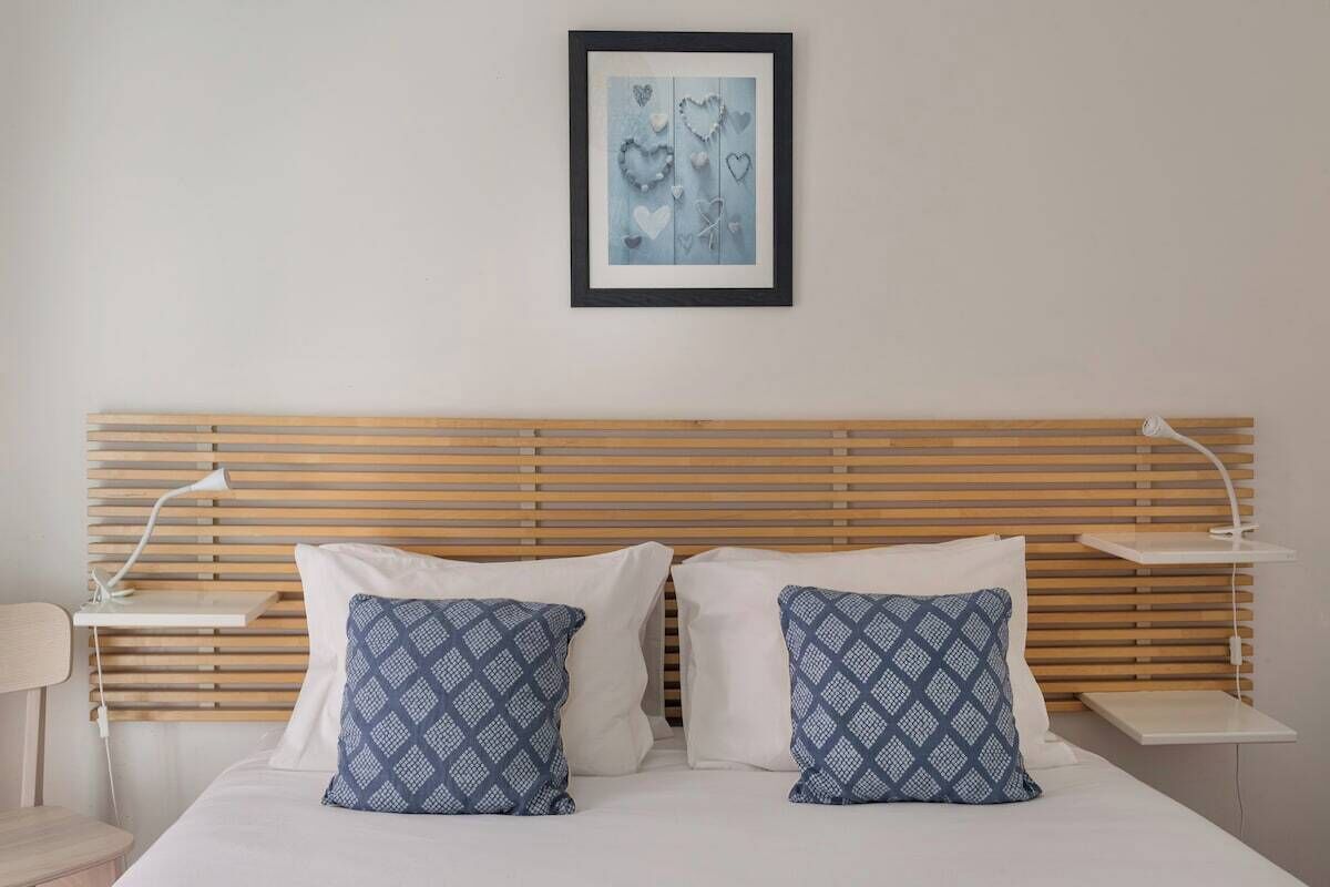 Una cama bien hecha con dos almohadas, un cabecero de madera, una mesita de noche con una lámpara y un cuadro enmarcado en la pared.