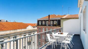 Uma varanda com mesa e cadeiras, com vista para uma paisagem urbana com telhados de terracota.