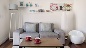 Een moderne woonkamer met een grijze bank, een bijzettafeltje met twee rode bekers, een vloerlamp en een witte stoel. Er zijn planken met ingekaderde foto’s en een grote poster van Lissabon.