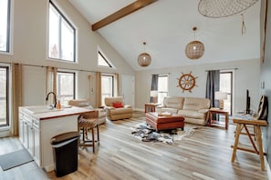 A spacious living room with a kitchen area, large windows, and a ceiling with exposed beams.
