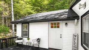 Une maison blanche avec un toit noir, une véranda avec un banc et des chaises, et une pancarte indiquant «Bienvenue».