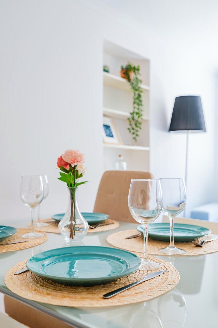 Una mesa de comedor con platos azul verdosos, un florero con una flor rosa y dos copas de vino.