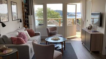 A modern living room with a sofa, armchairs, a coffee table, and a TV. There are large windows offering a view of the outdoors.