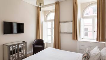 Une chambre à coucher comprenant un lit, une télévision, une chaise, une armoire et de grandes fenêtres avec des rideaux.