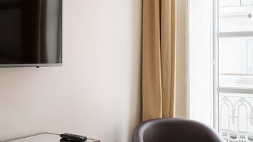 A room with a flat-screen TV mounted on the wall, a dark leather armchair, a wooden side table with a remote control, and a large window with a curtain.