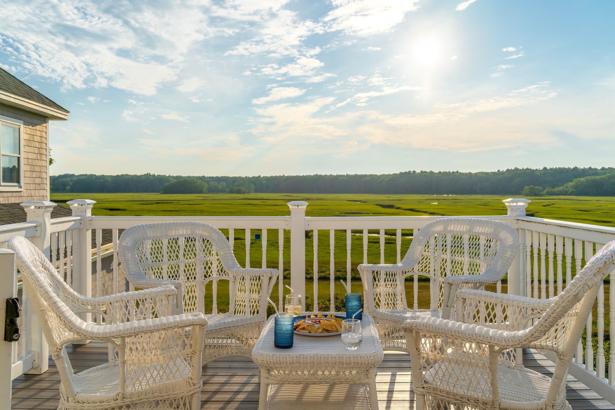 A deck with wicker chairs and a table, overlooking a green field and a house.