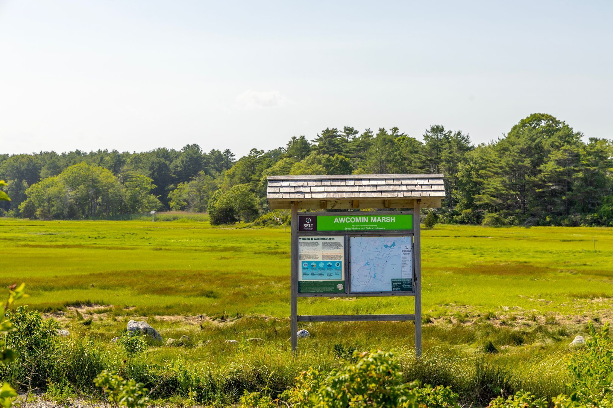 A signboard in a grassy field with text and maps, surrounded by trees.