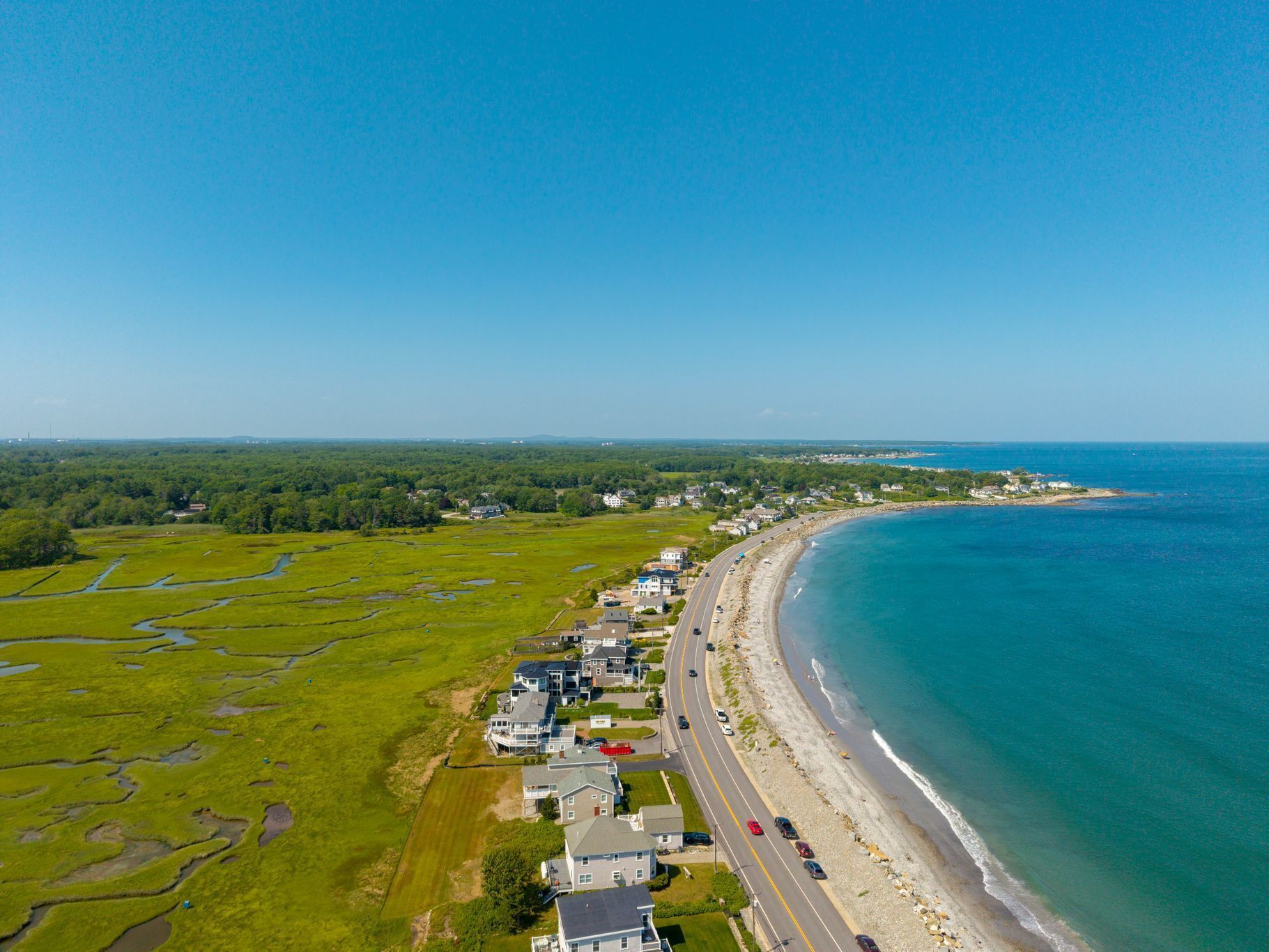 An aerial view of a coastal residential area with houses along a curved shoreline, a road parallel to the beach, and a clear blue sky.