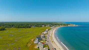 An aerial view of a coastal residential area with houses along a curved shoreline, a road parallel to the beach, and a clear blue sky.