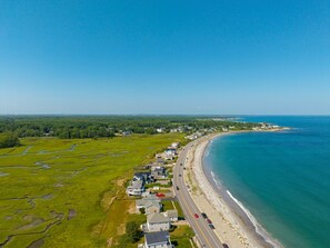 Luchtbeeld van een kustgebied met huizen langs een boogvormige kustlijn, een weg parallel aan het strand, en een helderblauwe lucht.