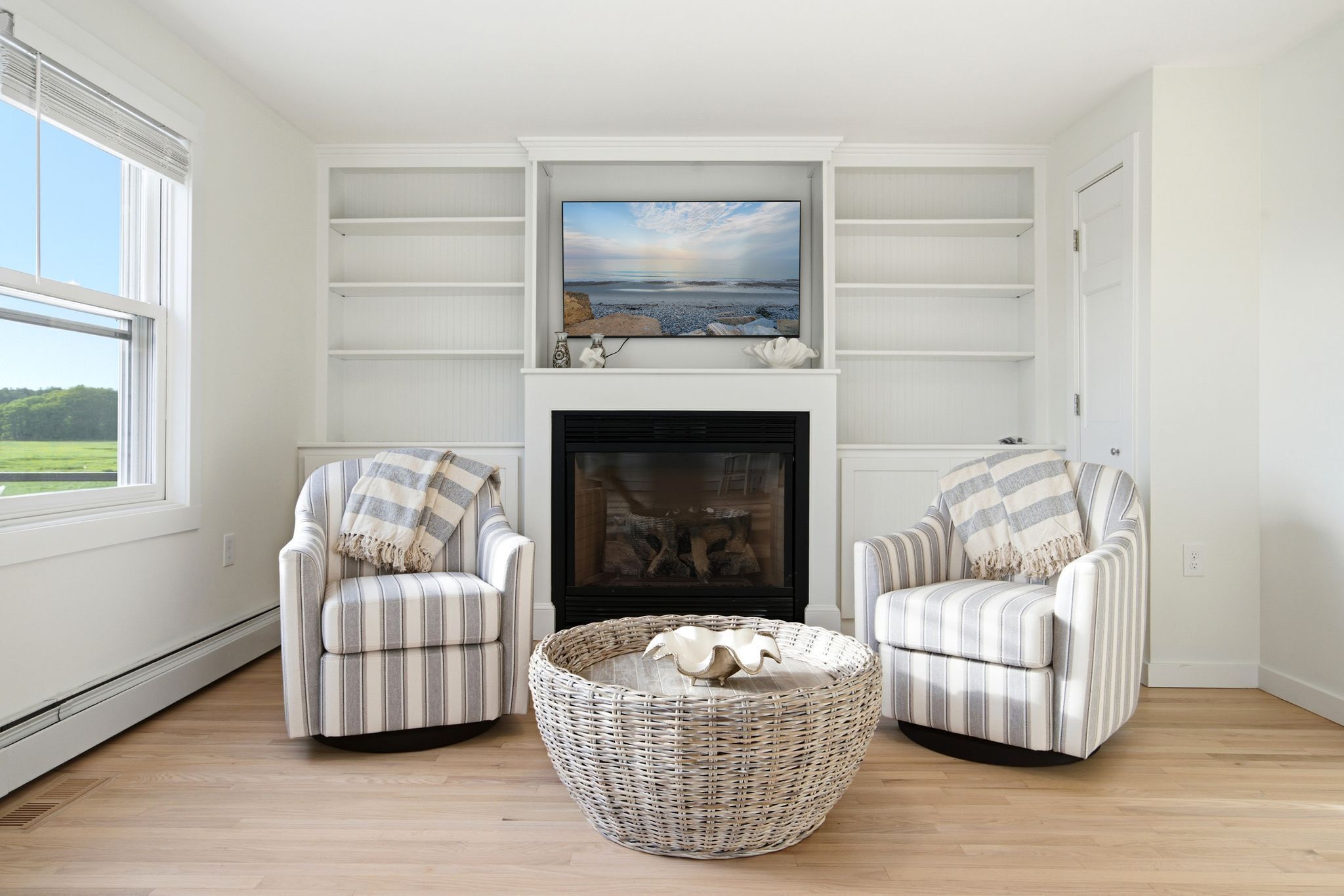 A living room with a fireplace, two striped armchairs, a woven ottoman, and a painting above the mantel.