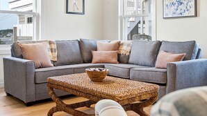 A grey sectional sofa with multiple cushions, a woven coffee table, and a framed bird painting on the wall.