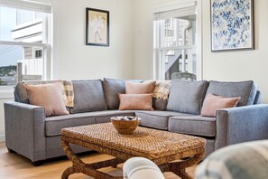 A grey sectional sofa with multiple cushions, a woven coffee table, and a framed bird painting on the wall.