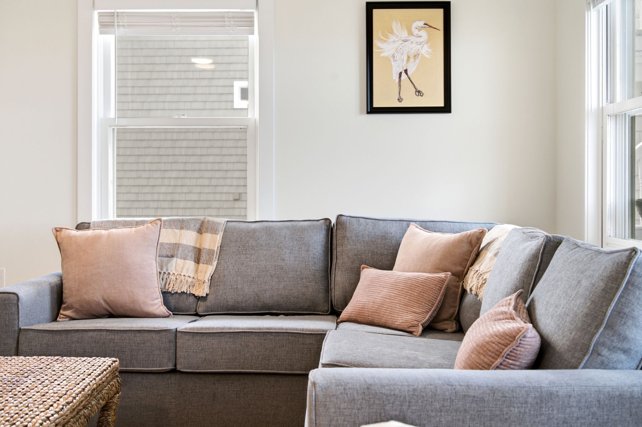A grey sofa with multiple cushions, a woven side table, and a framed artwork on the wall.