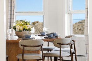 Een eetruimte met een tafel gedekt voor vier personen, stoelen, een bloemenvaas en uitzicht op het strand door het raam.