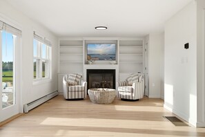 A spacious living room with a fireplace, two striped armchairs, a woven ottoman, and a large flat-screen TV mounted above the fireplace.