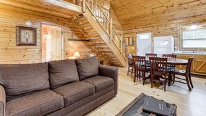 Een houten blokhut van binnen met een bank, een eettafel, stoelen, een trap en een koelkast.