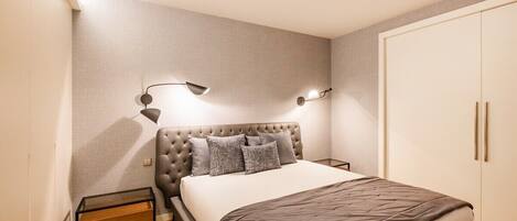 A bedroom with a tufted headboard, a bed with a white and brown bedspread, two bedside lamps, a wooden nightstand, and a wardrobe.