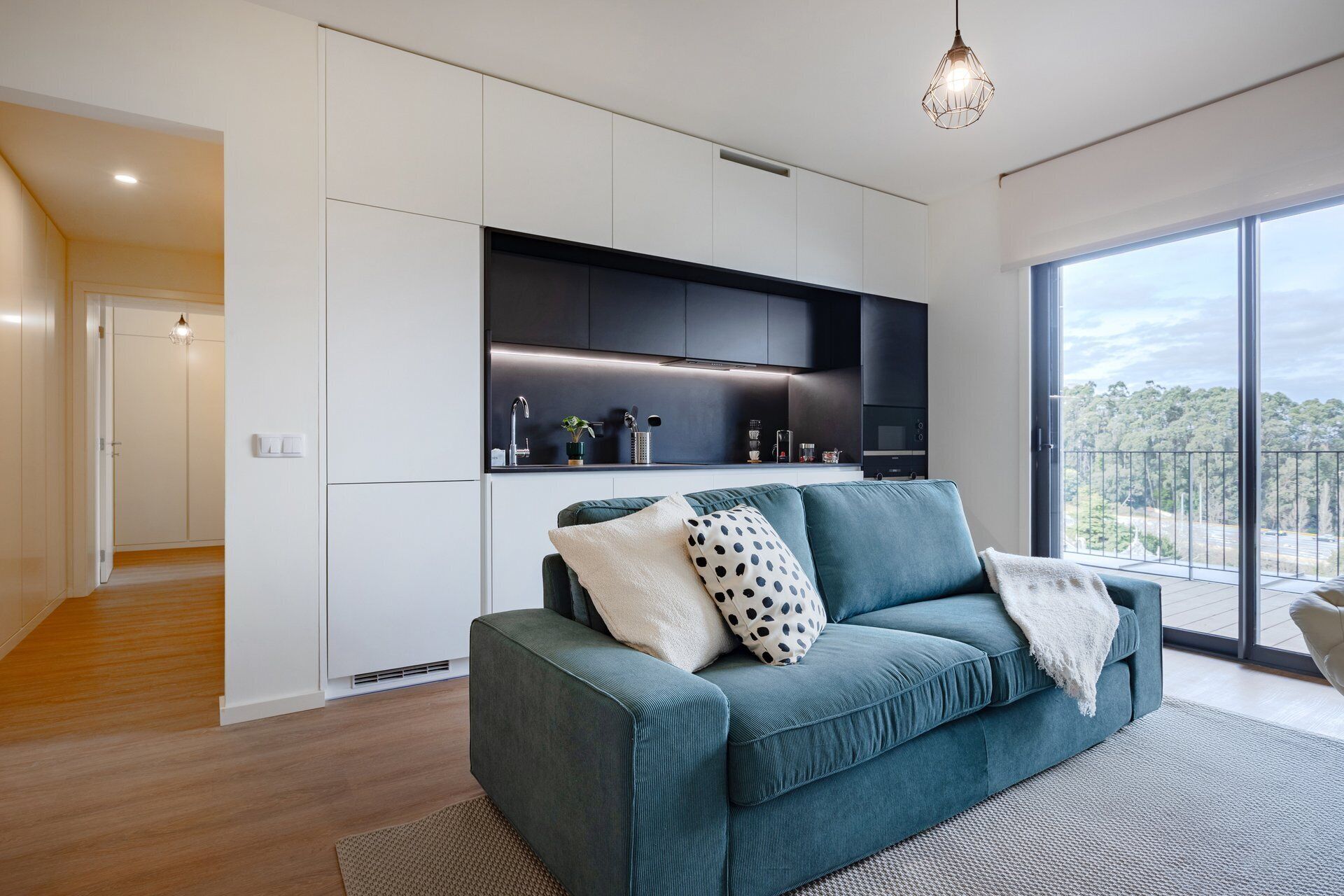 Un salon moderne avec un canapé de couleur turquoise, une cuisine dotée de meubles sombres et une grande fenêtre donnant sur la forêt.