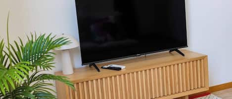 Un televisor sobre un pedestal de madera, con un control remoto, una lámpara blanca y una planta en maceta.