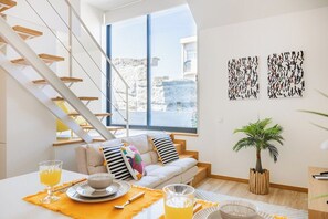 A modern living room with a staircase, a sofa, a dining table with orange placemats, and a potted plant. - Duque de Saldanha Duplex Loft (Porto)