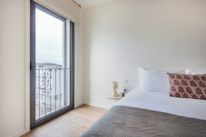 Ein Schlafzimmer mit großem Fenster, einem Bett mit weißen und gemusterten Kissen, einem Nachttisch mit Lampe und Blick auf Gebäude.