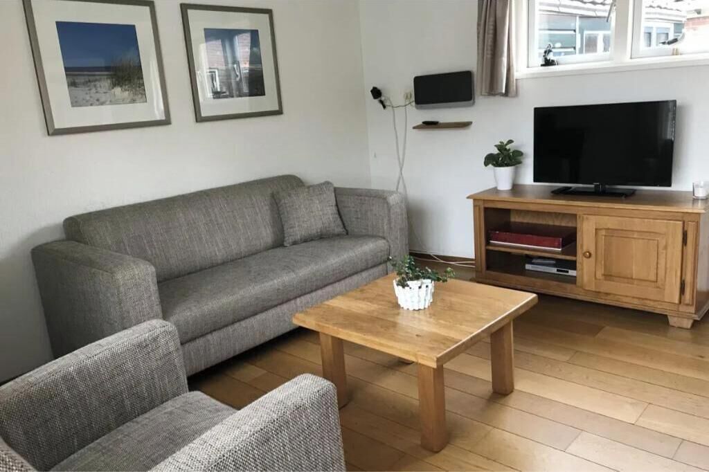 Uma sala de estar com um sofá cinzento, uma mesa de centro de madeira, uma televisão sobre um suporte de madeira e quadros nas paredes.