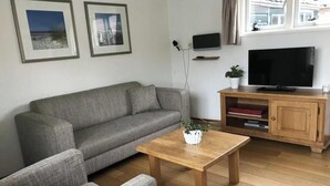 A living room with a grey sofa, a wooden coffee table, a television on a wooden stand, and framed pictures on the wall. - The Fisherman's Cottage (Egmond aan Zee)