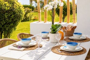 Un espace de restauration en plein air avec une table blanche dressée pour deux personnes, agrémentée de bols bleus et blancs, d’une carafe en verre et d’une plante en pot.