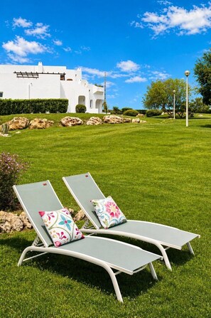Dos sillones de descanso con cojines estampados sobre un césped cuidado, un edificio blanco al fondo y un cielo azul despejado.
