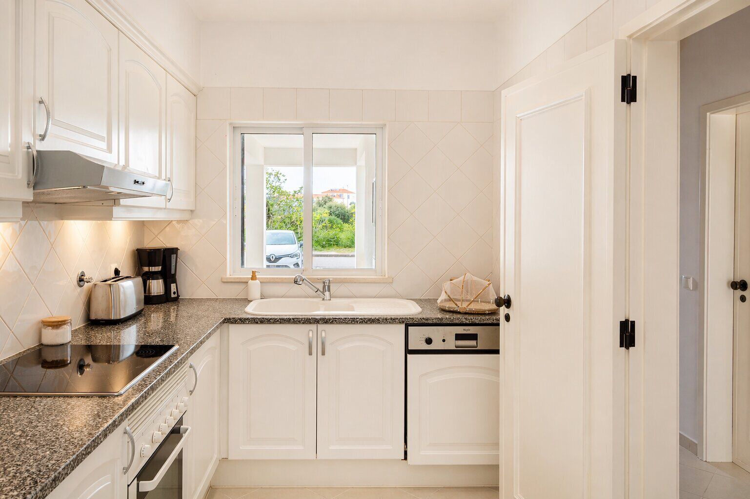 Eine moderne Küche mit weißen Schränken, einem Backofen, einem Kochfeld, einer Kaffeemaschine und einem Fenster mit Blick nach außen.