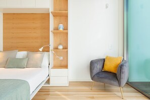 Une chambre à coucher moderne avec un lit, une lampe de chevet, des étagères en bois et un fauteuil gris agrémenté d’un coussin jaune.
