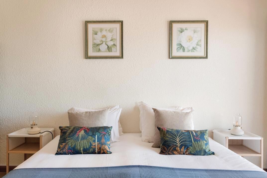 A neatly made bed with white linens and two decorative pillows, a bedside table with a lamp, and two framed floral artworks on the wall.