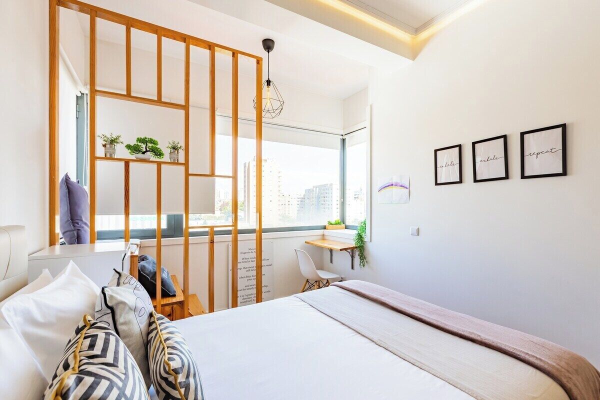 A modern bedroom with a large bed, a wooden partition, a desk with a chair, and a window with a city view.