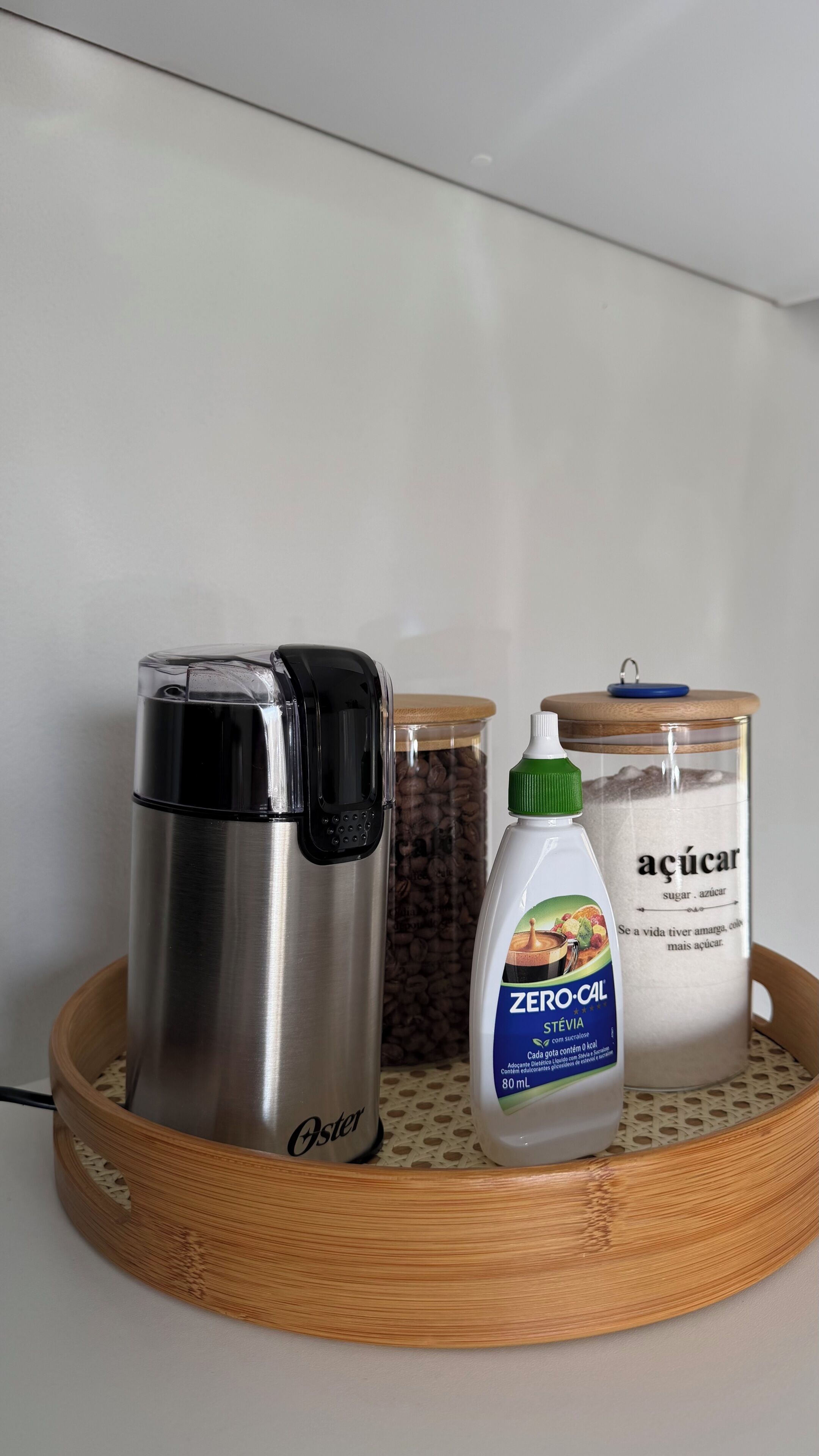 En stålfarvet Oster-kaffemaskine, en flaske Zero-cal Stevia, og en glasdåse mærket 'açúcar' på en træbakke.