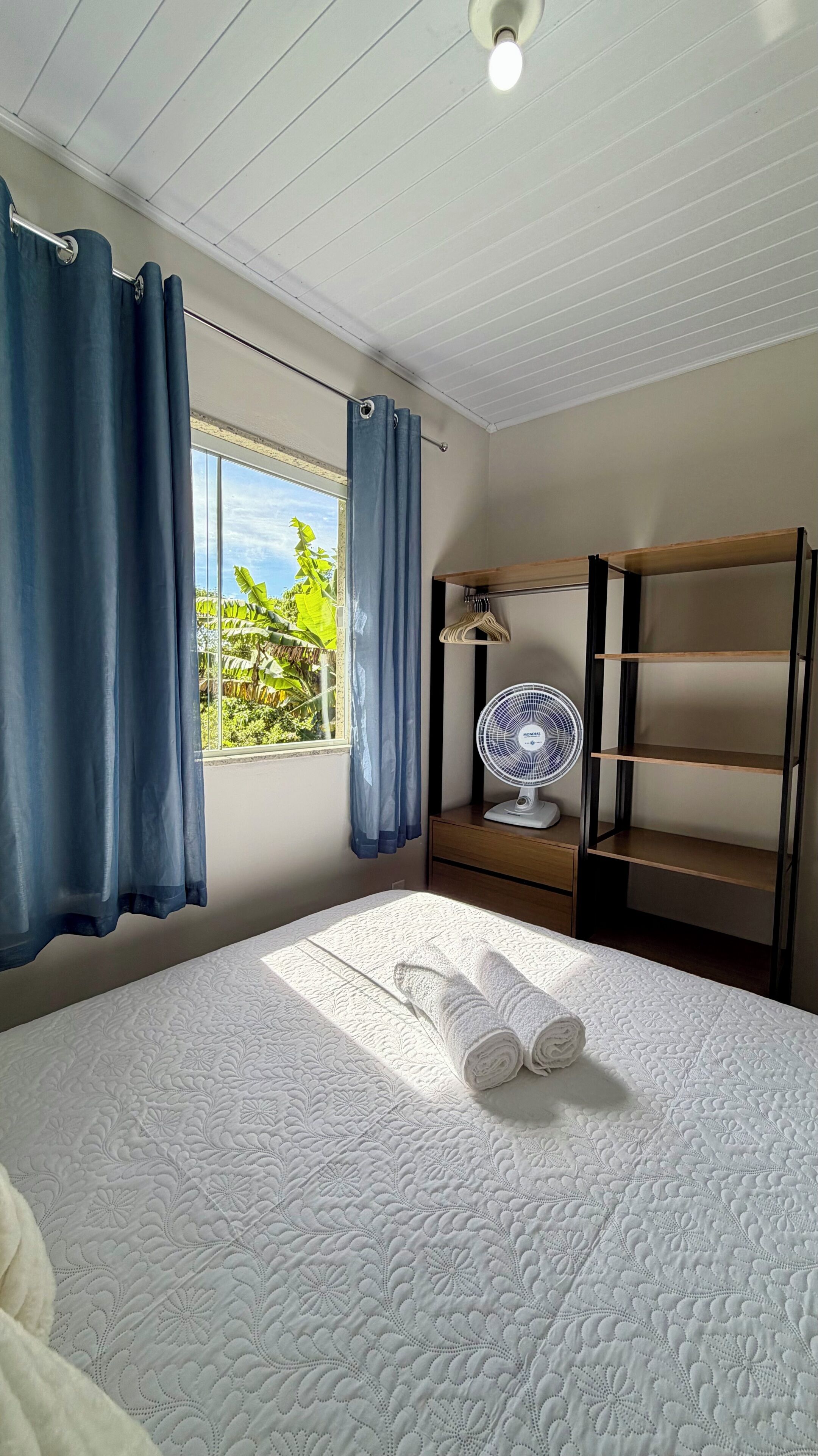 A bedroom with a bed, a window with curtains, a fan, and a shelf with hangers.