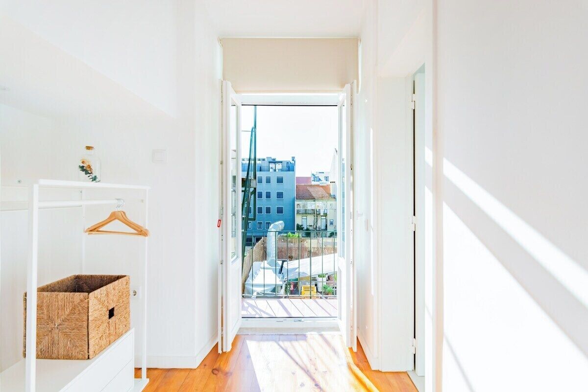 A narrow hallway with a wooden floor, a white door, and a view of buildings through an open door.