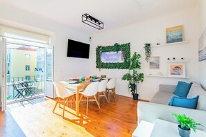 A bright living room with a wooden floor, a dining area with a table and chairs, a sofa, a TV mounted on the wall, and a balcony with a view of buildings. - Superior Suite @ Guest Inn 1 Boutique House (campo pequeno)