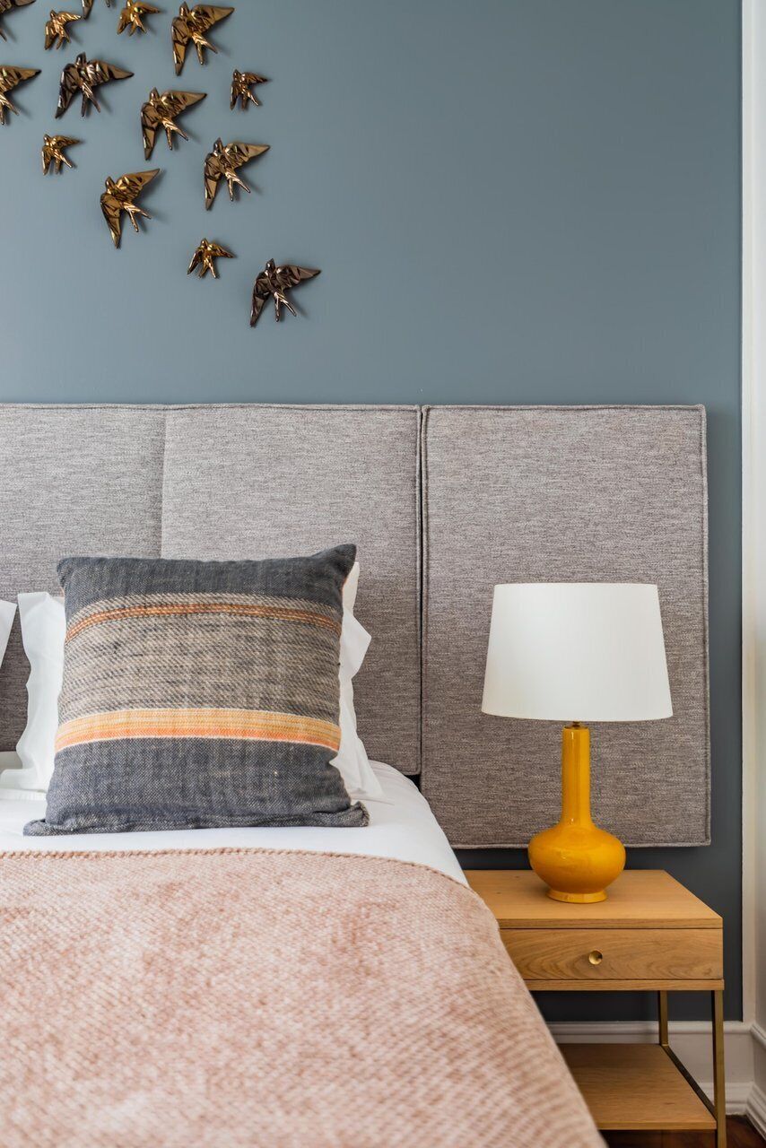 A bedroom with a grey headboard, a patterned pillow, a bedside lamp, and a wall decoration.