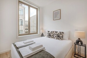 Habitación de hotel con cama, dos lámparas de noche, una ventana con vistas a edificios y un cuadro en la pared.