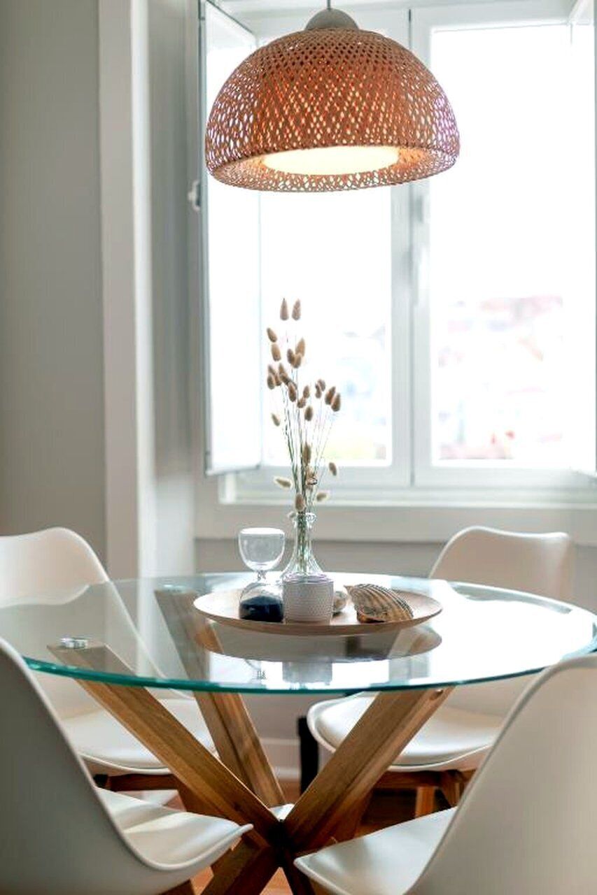 A round glass table with a woven pendant light, white chairs, and a vase with dried plants.