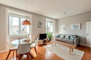 Een moderne woonkamer met een glazen eettafel, een bank, een televisie en grote ramen.