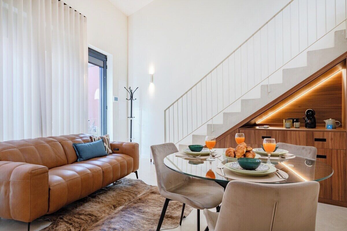 A modern living room with a leather sofa, a glass dining table set for breakfast, and a staircase with a wooden finish.