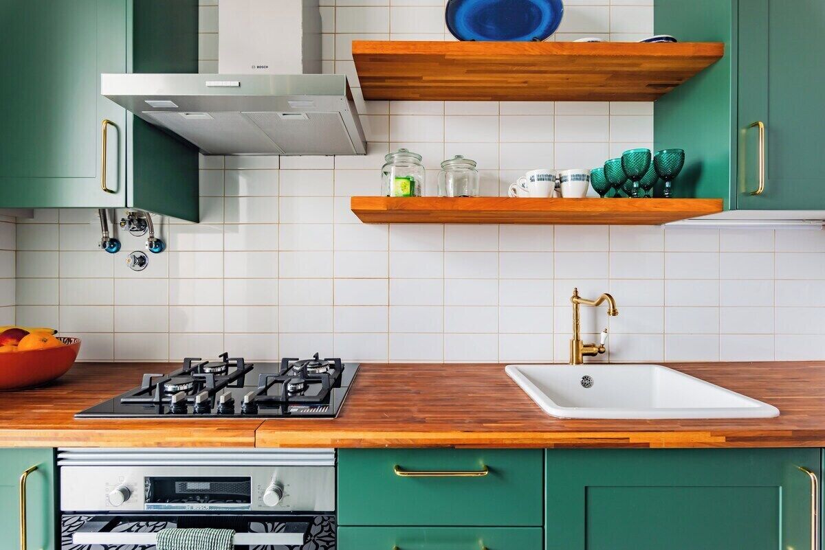 A kitchen with green cabinets, a gas stove, a white sink, and wooden shelves.