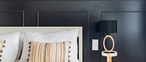 A neatly made bed with white bedding and patterned pillows, a black-framed picture on the wall, a black lamp with a wooden base, and a small wooden stool with a white tray.