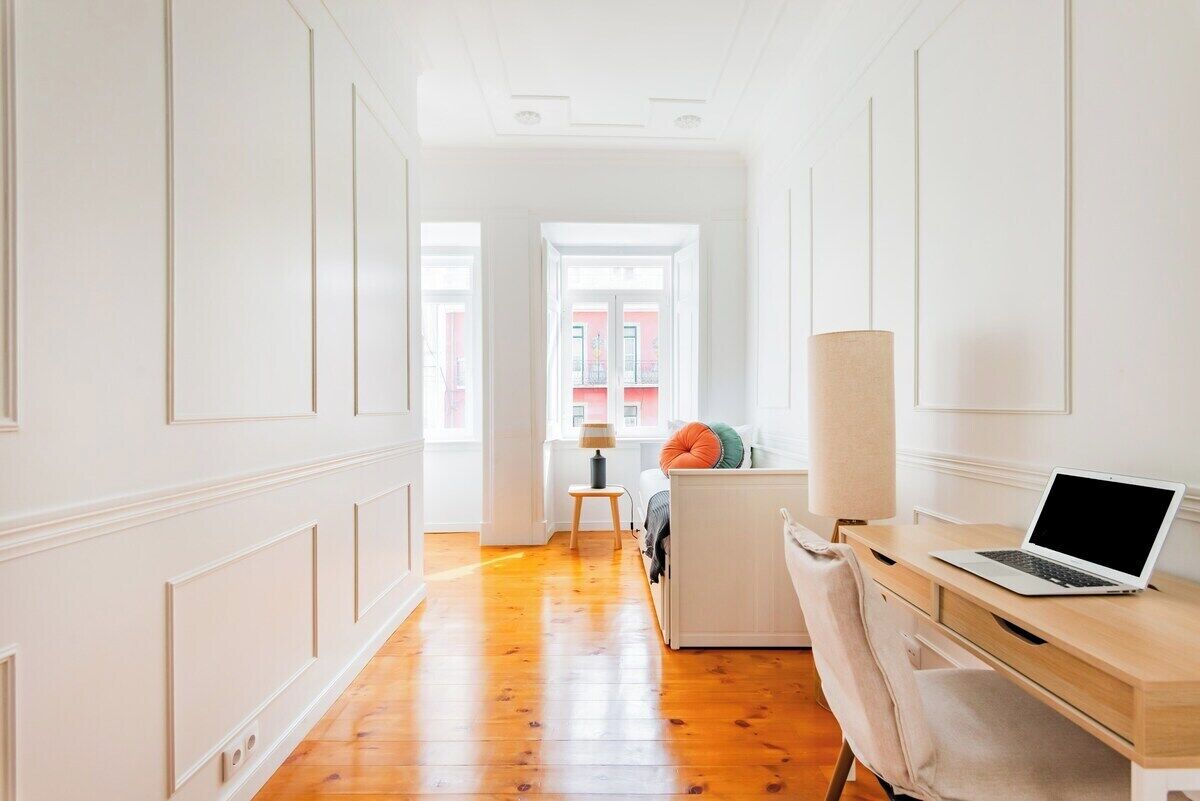 A hallway with a wooden floor, a desk with a laptop, a lamp, and a bed with pillows.