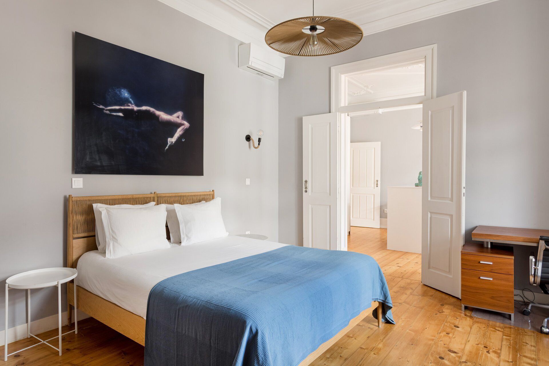 Een slaapkamer met een bed, een blauwe deken, een houten hoofdbord, een wandlamp en een groot schilderij.