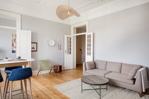 Una sala de estar con un sofá color beige, una mesa redonda de centro, suelo de madera y una puerta blanca que da a otra habitación.