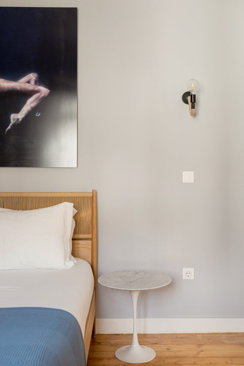 A bedroom with a bed, a small round table, a wall-mounted light, and a painting.